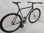 TrackloX Midnight Mirage TLX20MM – a matte black single-speed fixie bike with a chrome fork, purple hubs, and Arisun tires. Perfect for urban riding, bikepacking, and tracklocross. Captured from a rear-side angle