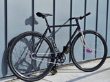 TrackloX Midnight Mirage TLX20MM – a matte black single-speed fixie bike with a chrome fork and purple hubs, leaning against a modern metal wall. Built for urban riding, bikepacking, and tracklocross