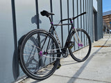 TrackloX Midnight Mirage TLX20MM – a black single-speed fixie bike with a chrome fork, purple hubs, and Arisun tires, leaning against a modern blue wall near an urban transit station. Built for urban riding, bikepacking, and tracklocross