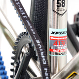 Close-up of the XFIXXI TrackloX Urban Bike (Single-Speed Edition) frame sticker on the central tube. Featuring a stainless steel frame with 'Engineered in Canada' and '4130 Chromoly' decals, alongside the Gates Carbon Belt Drive system