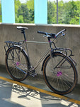 XFIXXI TrackloX Touring Bike with Gates Carbon Belt Drive, front and rear racks, and 35c gravel tires. Photographed in an urban setting with natural lighting, showcasing its versatility for city commuting and long-distance touring